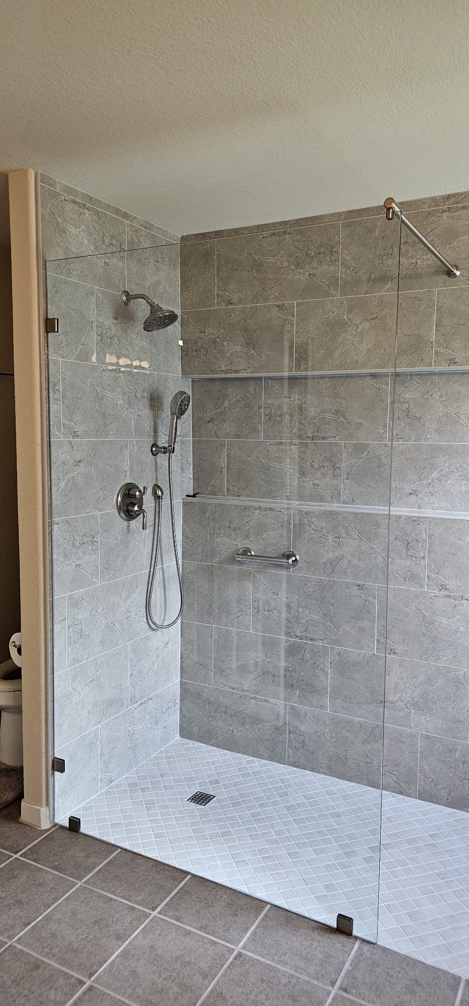 Glas Plus wide fixed panel reference photo: a wide fixed glass panel, no door, in an open grey tile shower, with a diagonal support bar at the top