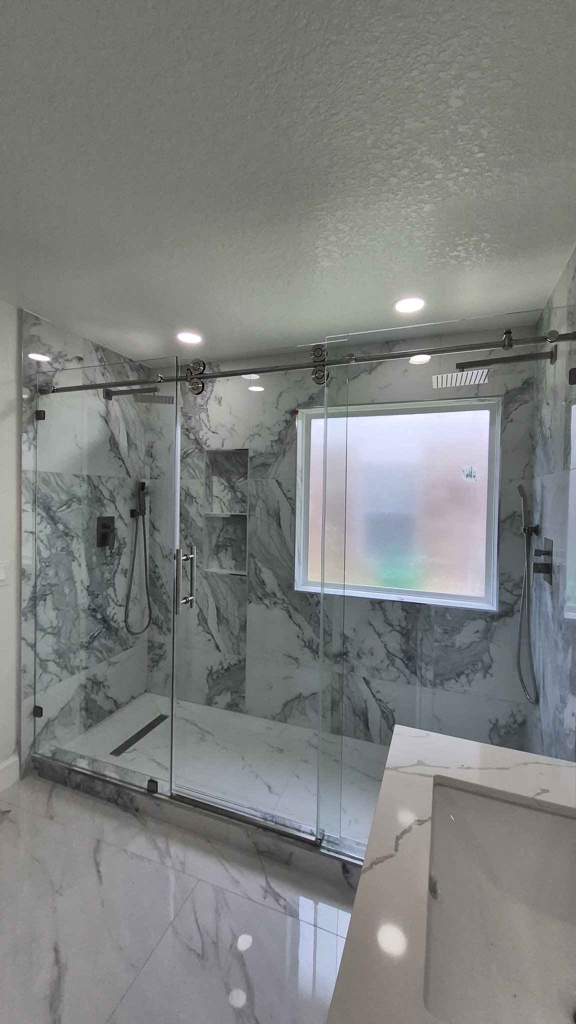 Wide clear glass sliding door with a chrome track in a large marble shower with an interior window