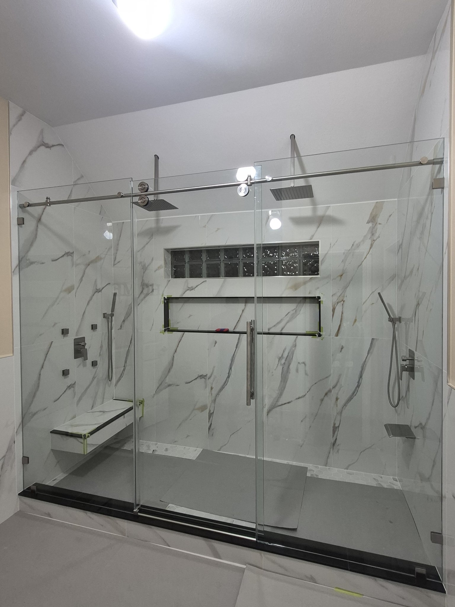 Sliding with center door (fixed panel, door and fixed panel) with a brush nickel track in a large marble shower with two showers and a bench